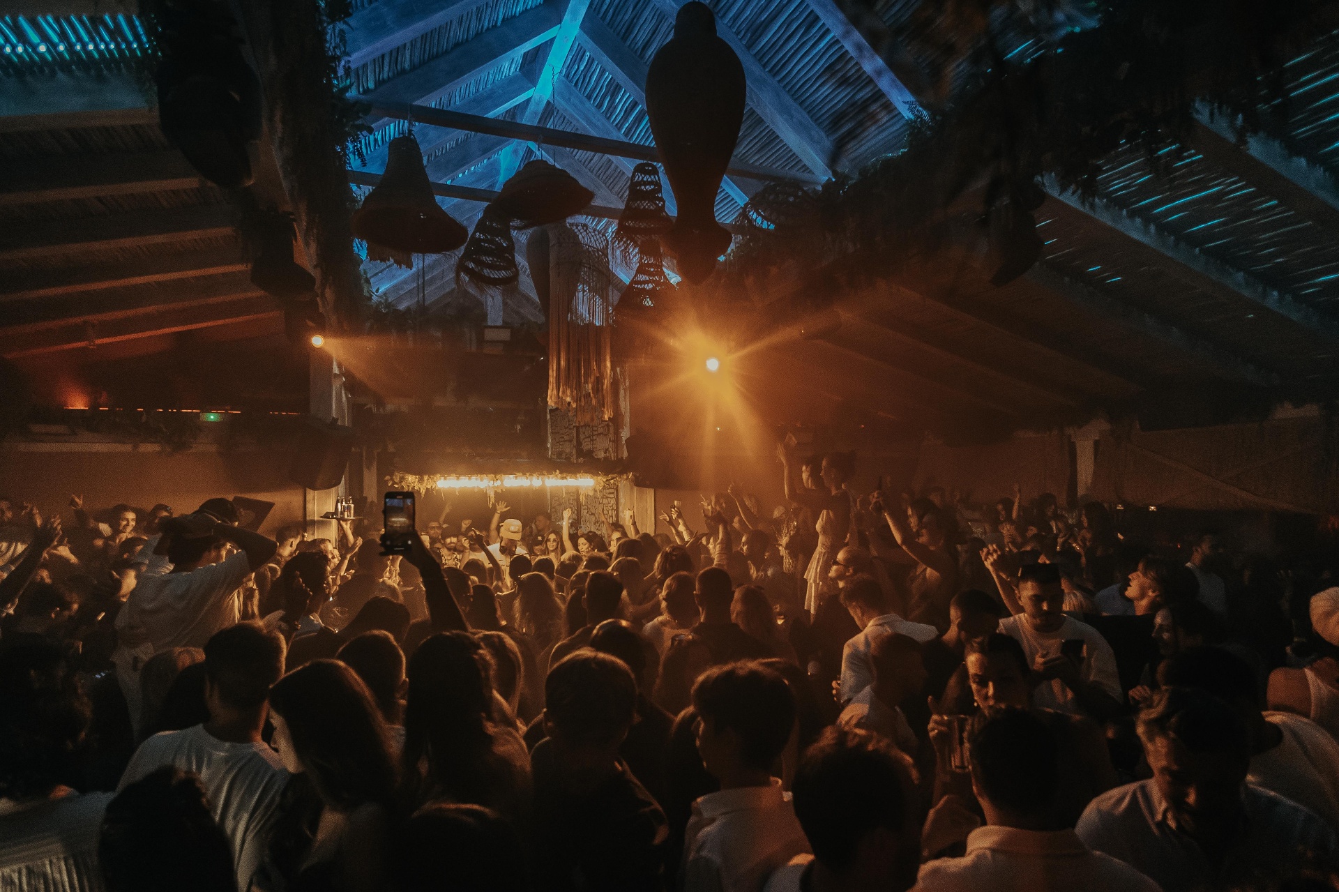 Packed night-time dance floor under a thatched roof at a Marbella beach club, with warm stage lights, raised hands and an energetic party crowd dressed in white